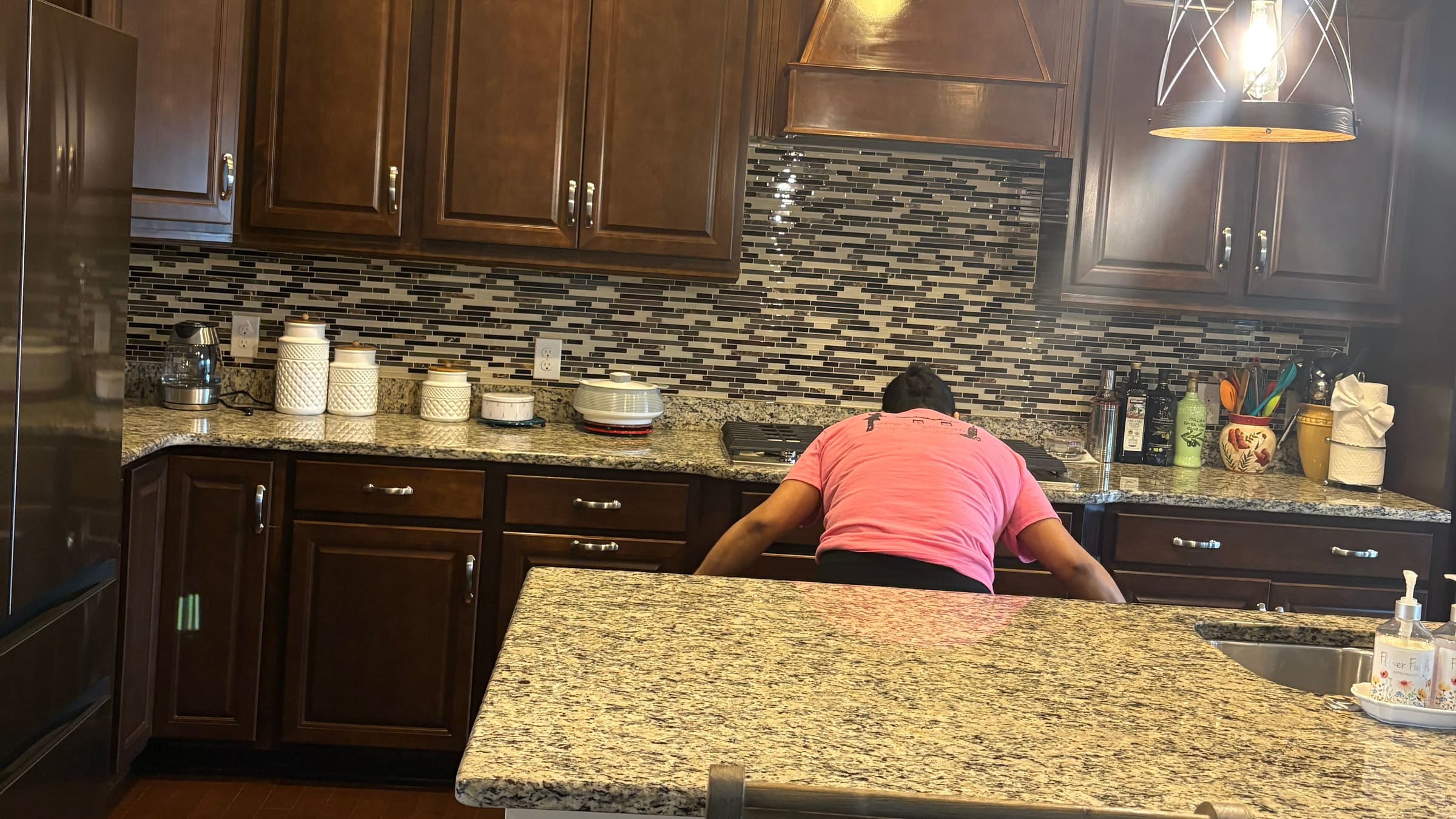 One of Rossy's cleaners in a pink shirt reaching across a granite worktop to wipe it down, in a client's kitchen.