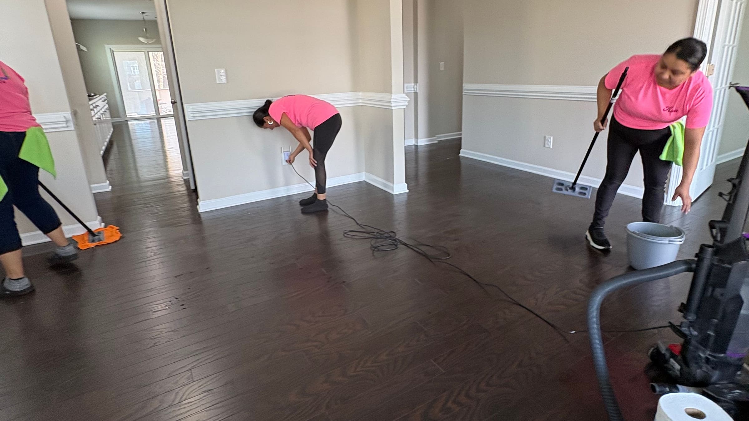 Three of Rossy's cleaners in pink shirts working a Charlotte dining room: one mopping the hardwood, one wiping a baseboard, one running a vacuum.